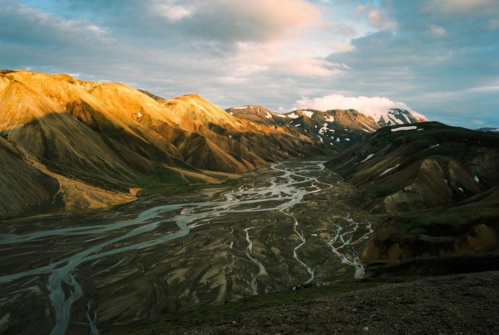 A wide river with many tributaries flows through a valley surrounded by mountains, some of which are covered in snow.