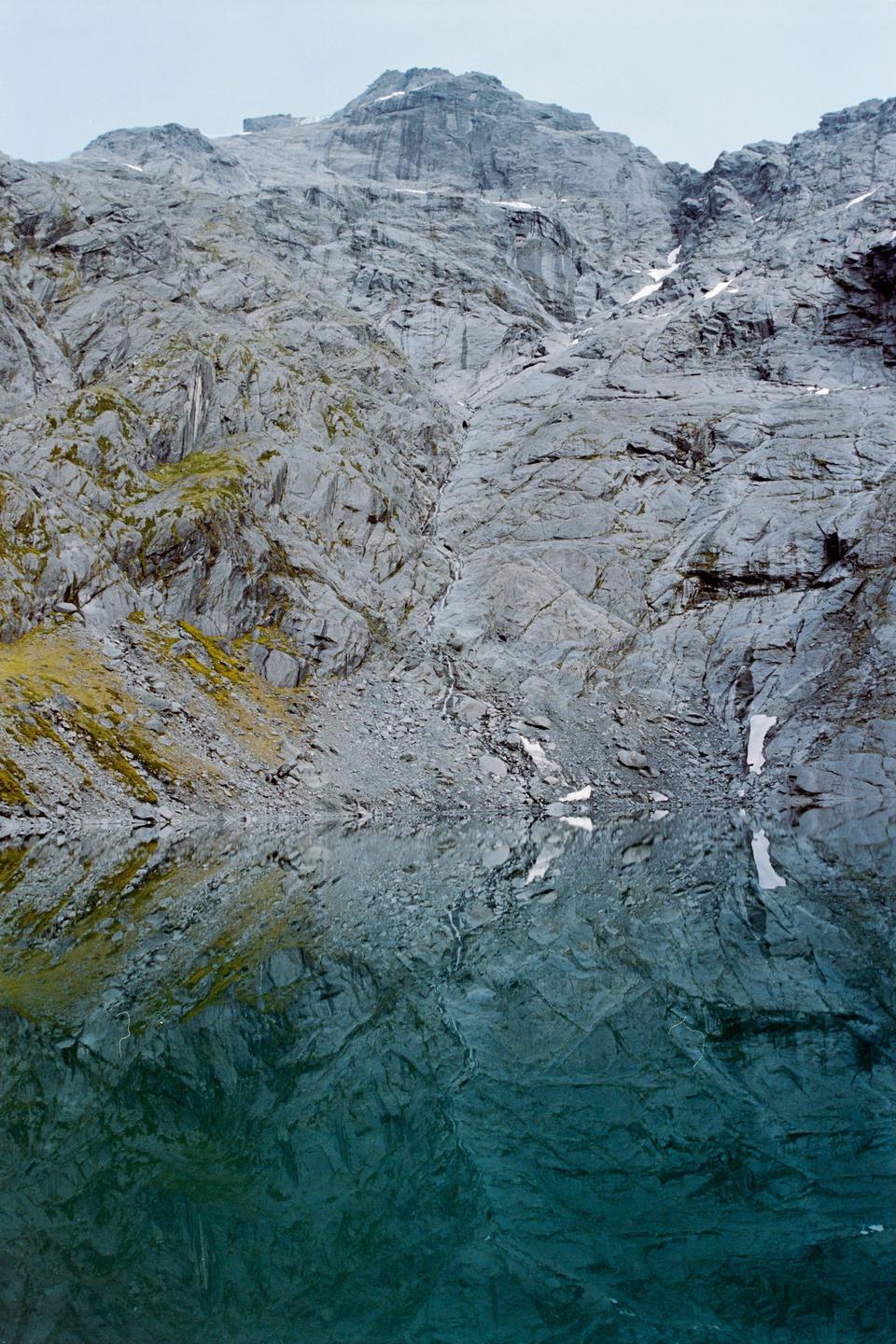 A mountain with patches of snow and moss is reflected in a still lake.