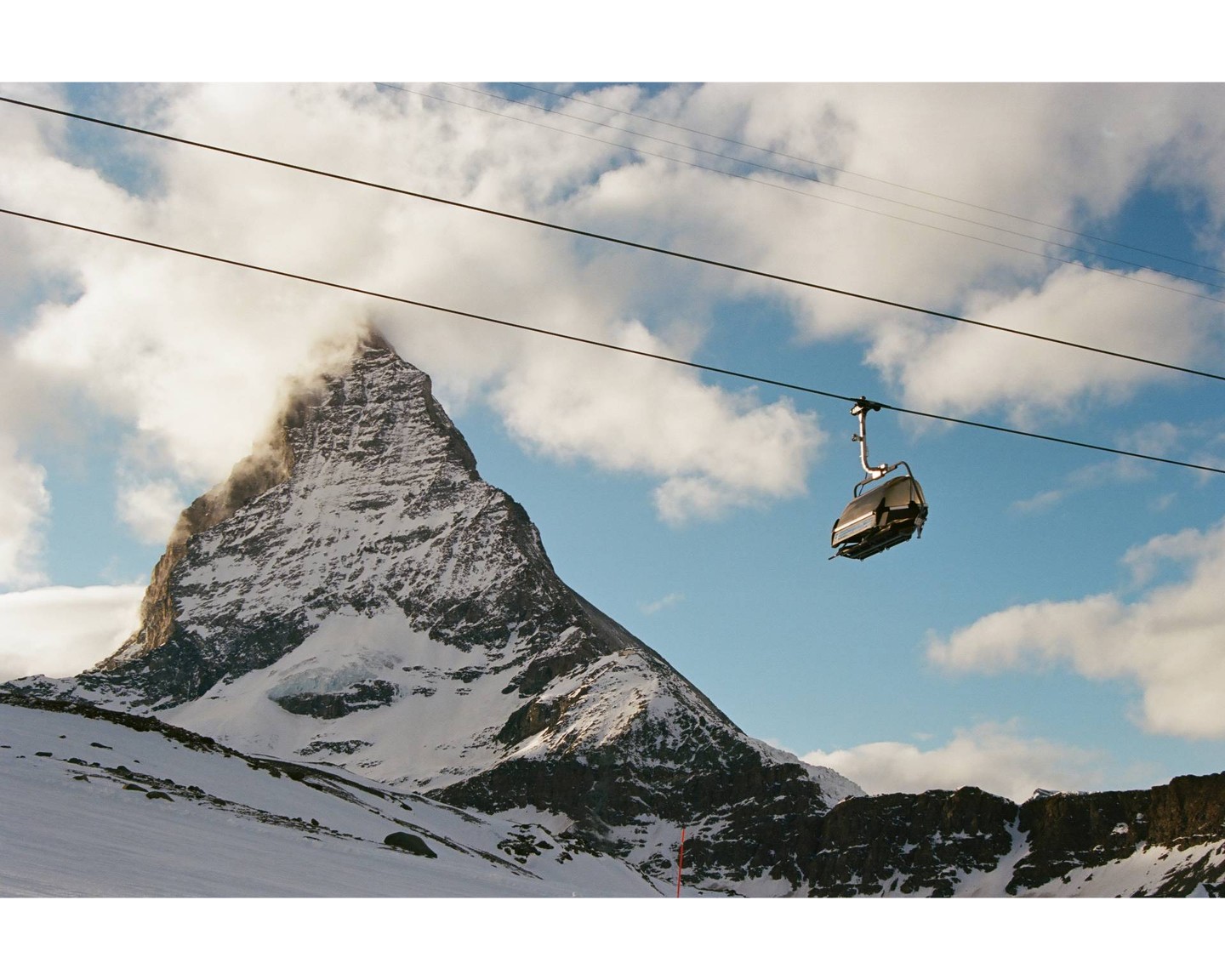 A ski lift chair hangs from cables above a snow-covered mountain peak partially obscured by clouds.