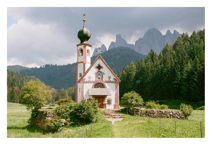 A white and red church with a domed steeple stands in a green field with mountains and trees in the background.