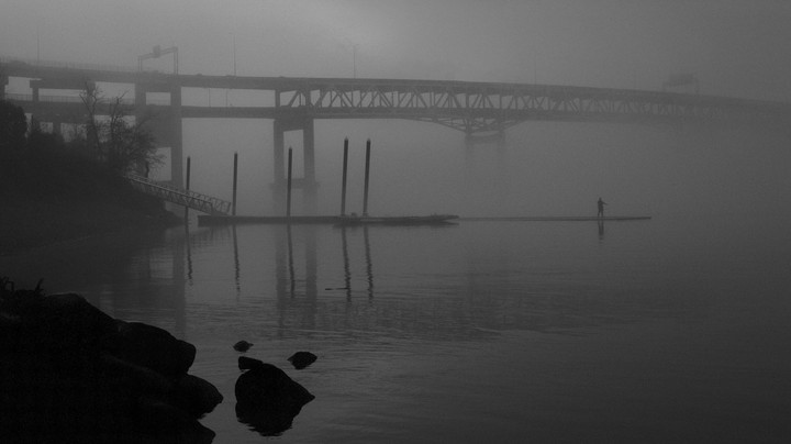 A lone figure stands on a floating dock in a foggy river with a bridge in the background.