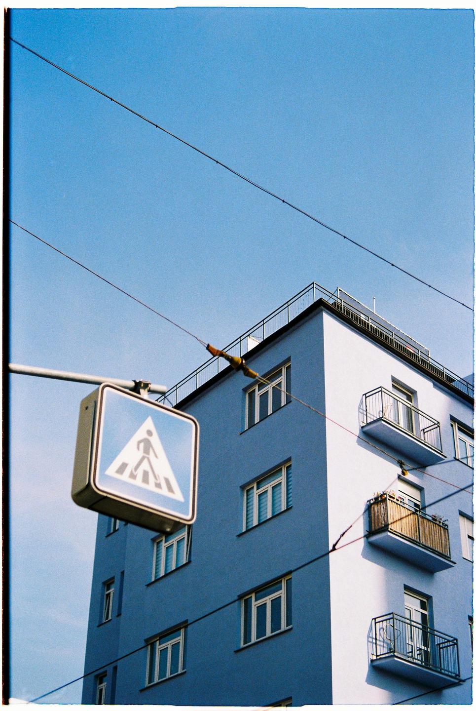 A pedestrian crossing sign hangs in front of a blue building with balconies and power lines crossing the frame.