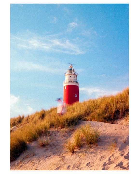 A red and white lighthouse stands on a sandy dune covered with dry grass under a blue sky.