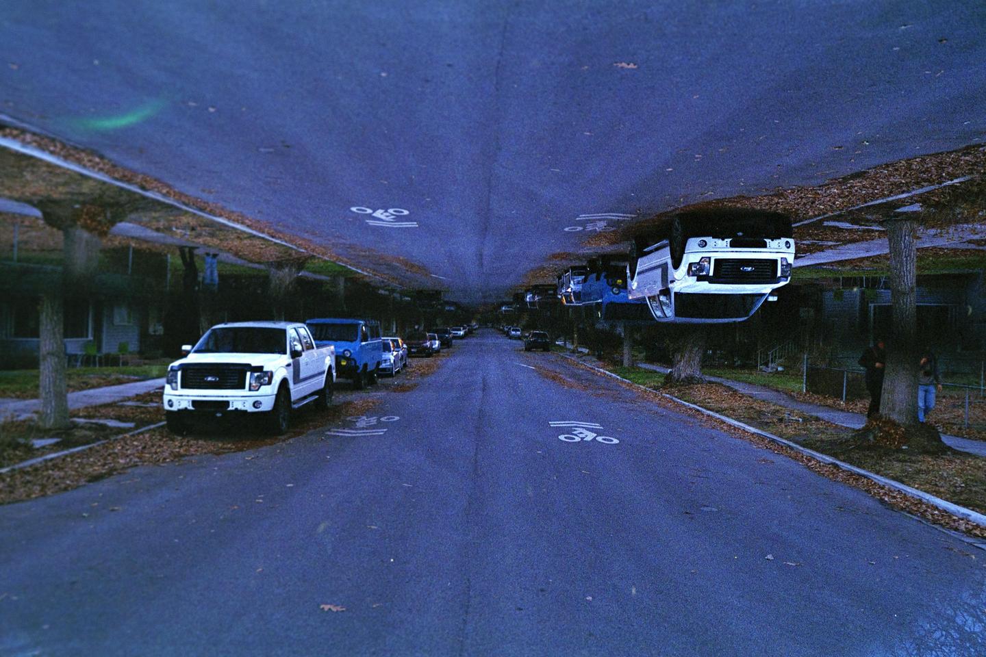 A street with parked cars is shown with an inverted, mirrored image of the same street above it, creating a surreal effect.