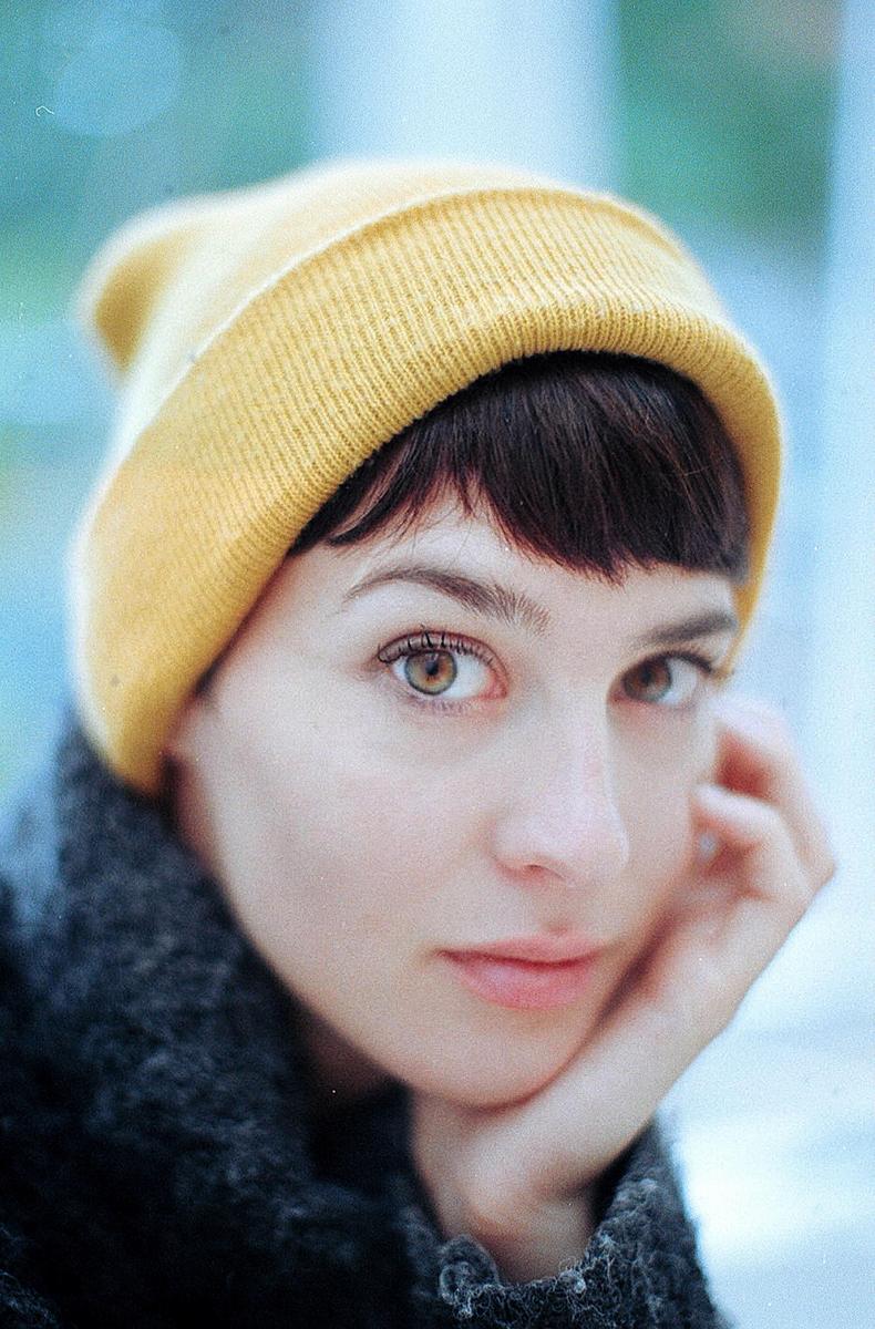 A woman wearing a yellow beanie looks directly at the viewer, resting her chin on her hand.