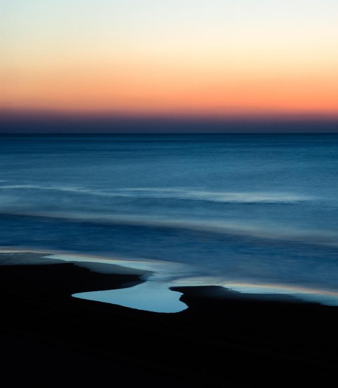 The ocean meets a dark beach with a small pool of water under a colorful sunset sky.
