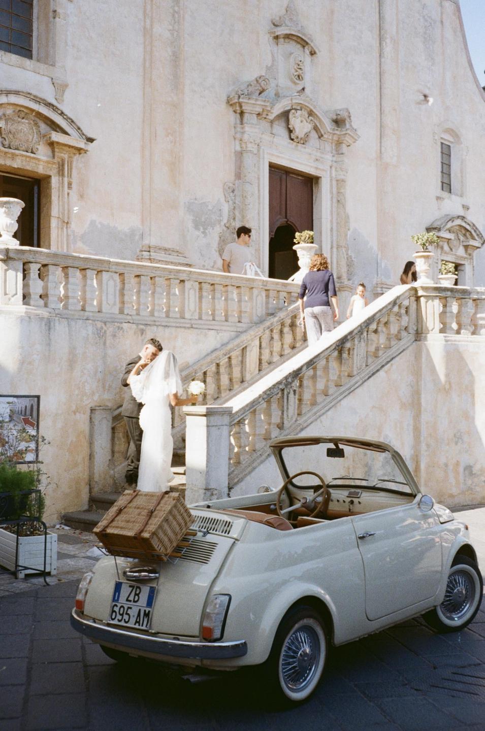 A couple stands on a stone staircase next to a small white convertible car with a wicker basket on the back.
