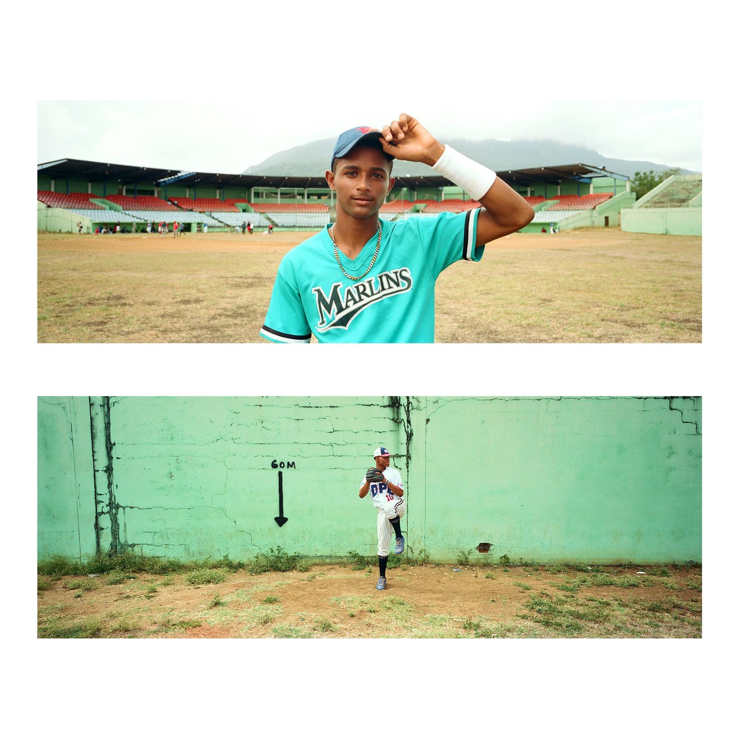 A young man in a baseball uniform stands on a dirt field with a stadium in the background, and another young man in a baseball uniform pitches in front of a green wall.