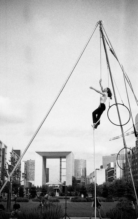 A woman performs acrobatics on a rope in an urban park with modern buildings in the background.