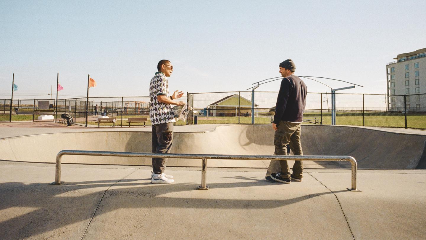 Two men stand in a skatepark, one holding a skateboard, with a building and flags visible in the background.