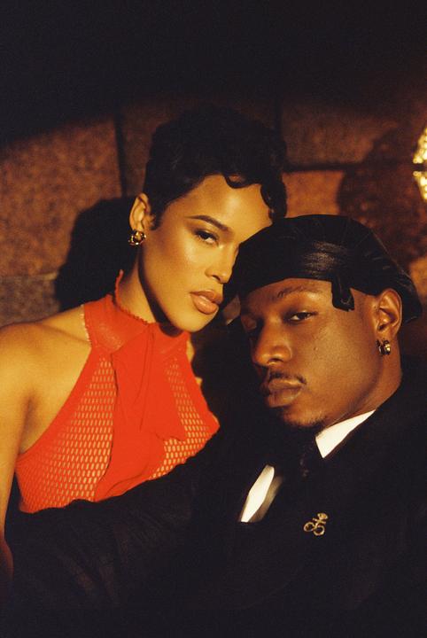 A woman in a red top and a man in a black suit and durag pose for a portrait.