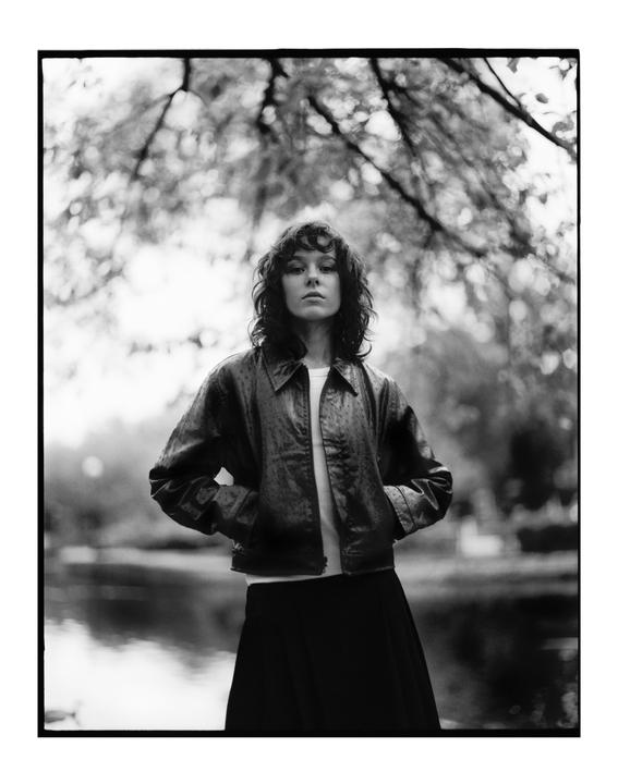 A woman with curly hair stands with her hands in her pockets, with a body of water and trees in the background.