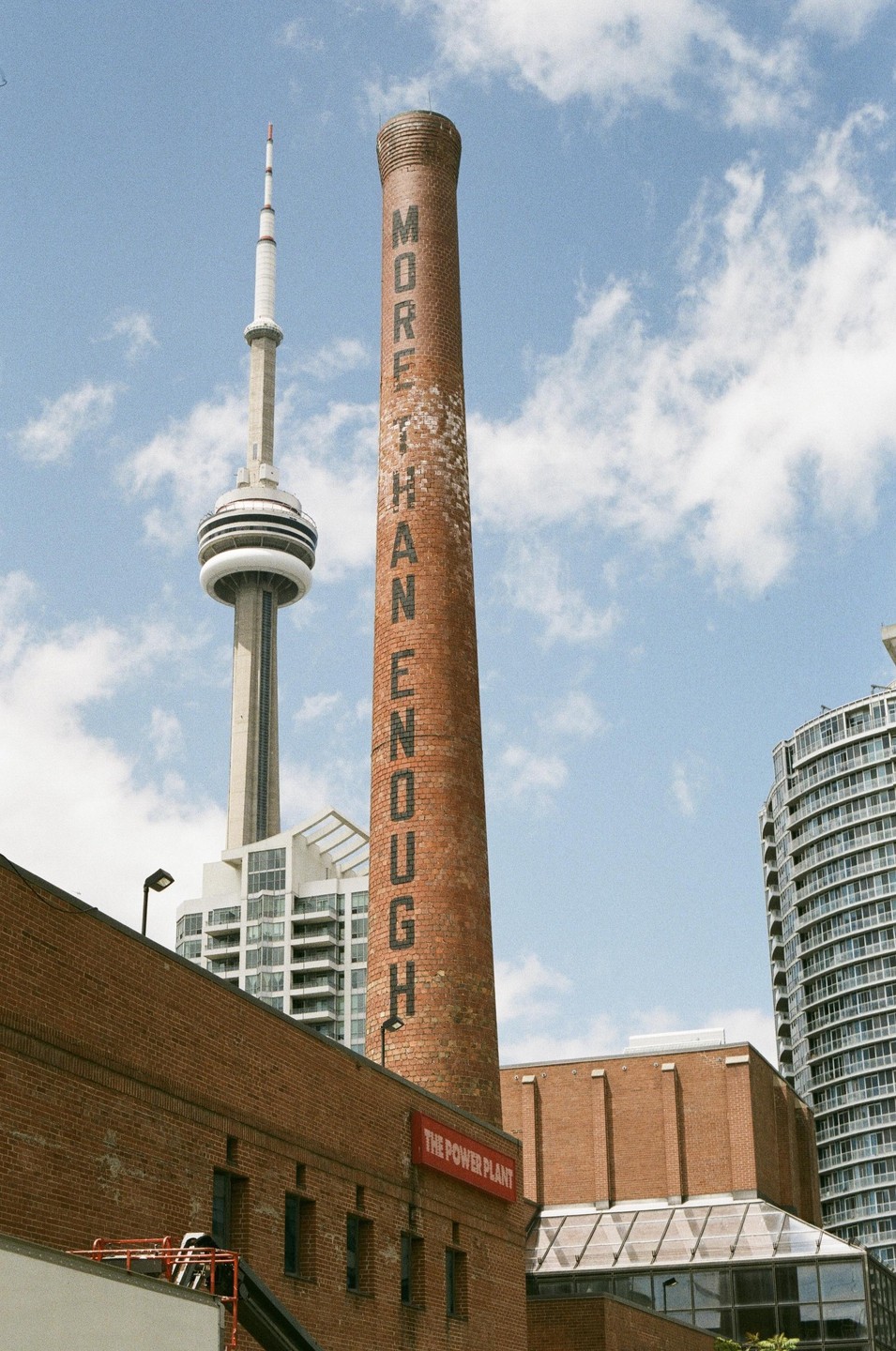 A tall brick chimney with "MORE THAN ENOUGH" written on it stands next to the CN Tower and other buildings under a cloudy sky.