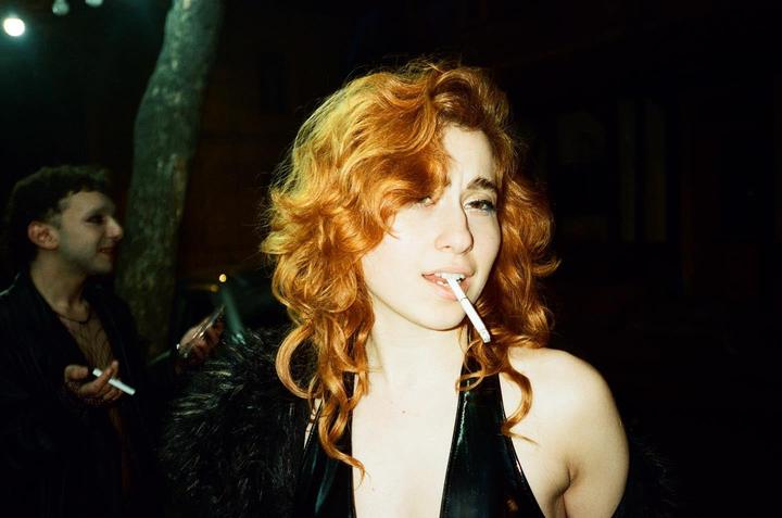 A woman with red hair and a shiny black top holds a cigarette in her mouth while a man in dark clothing stands behind her.