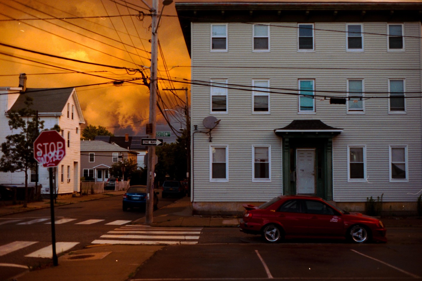 A red car is parked on a street corner with a stop sign and a building with many windows under an orange sky.