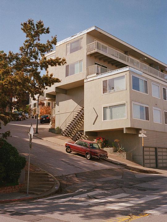 A red car is parked on a steep street next to a multi-story building with external staircases.