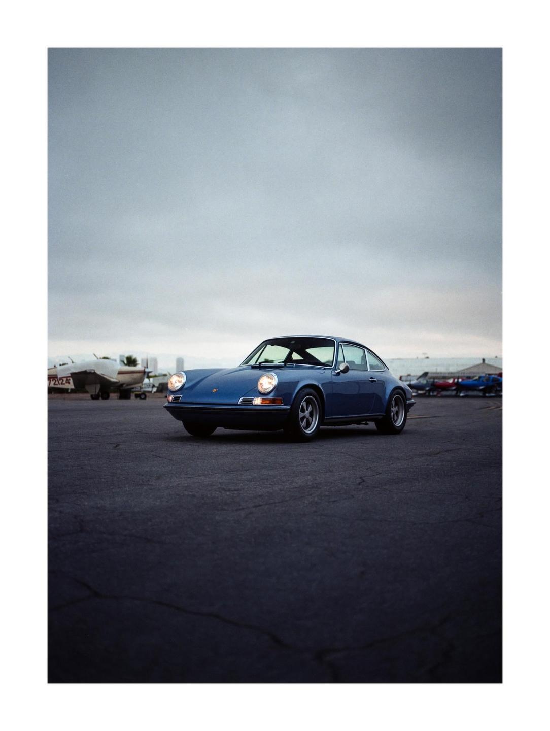A blue car is parked on an asphalt surface with airplanes in the background under a cloudy sky.