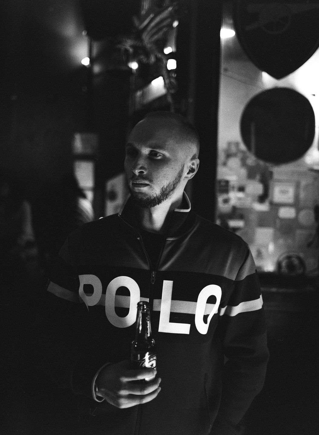 A man with a beard holds a beer bottle and wears a jacket with "POLO" written on it.