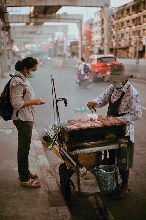 A woman wearing a face mask stands next to a street vendor grilling food on a cart.