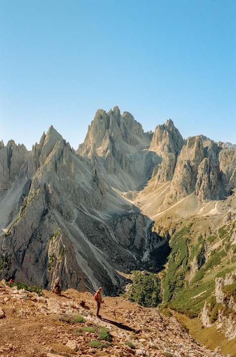 A person with a backpack and hiking poles stands on a rocky mountain path overlooking a valley with jagged peaks under a clear sky.