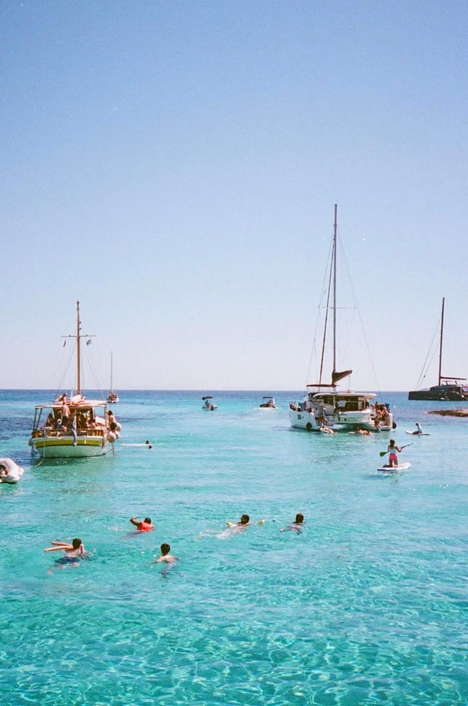 People swim and paddleboard in clear blue water near several boats under a bright sky.