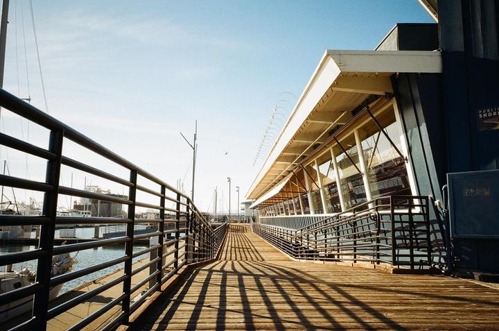 A wooden pier with metal railings runs alongside a building with large windows, with boats visible in the water beyond.
