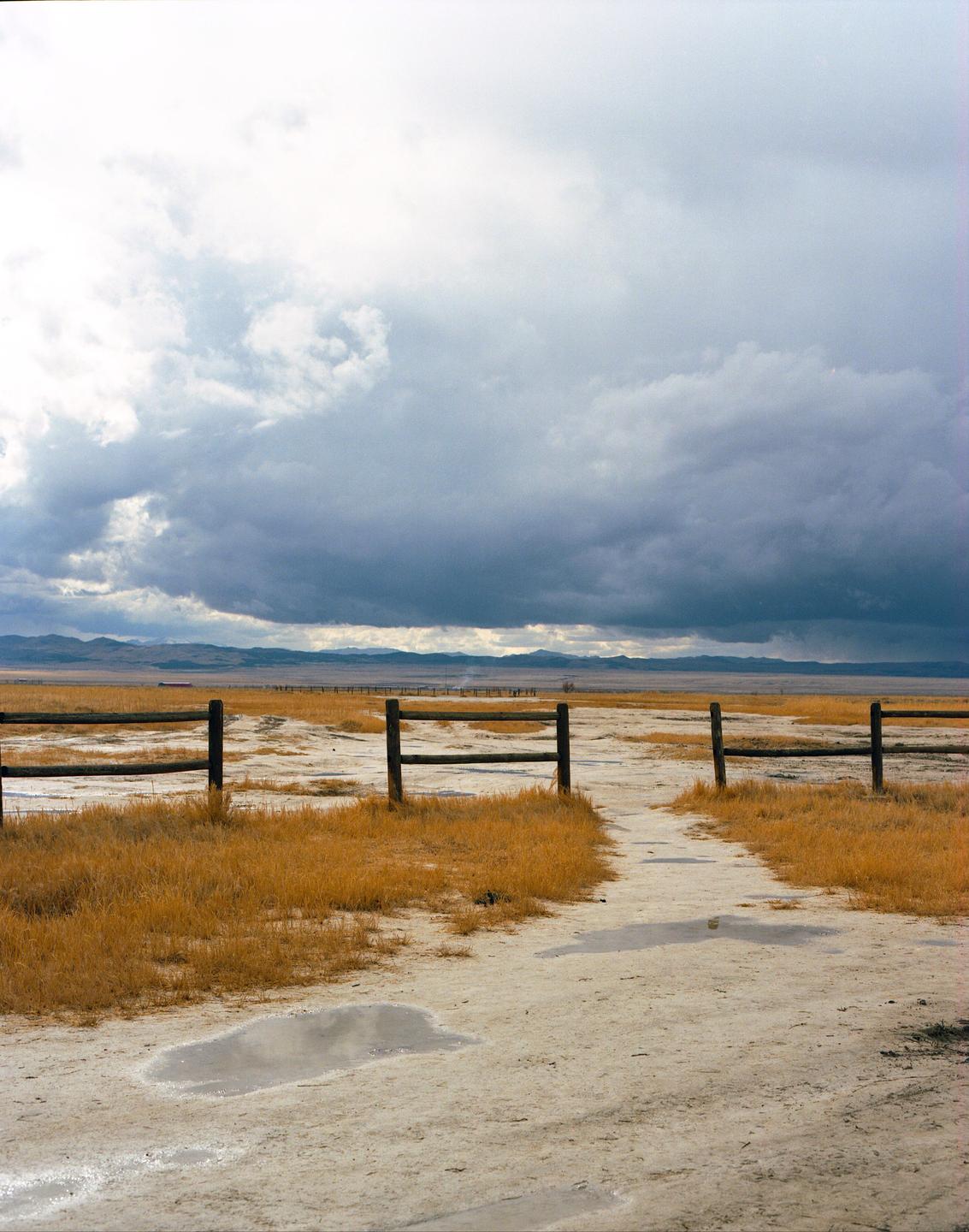 A dirt path leads through a wooden fence into a field of dry grass under a cloudy sky.