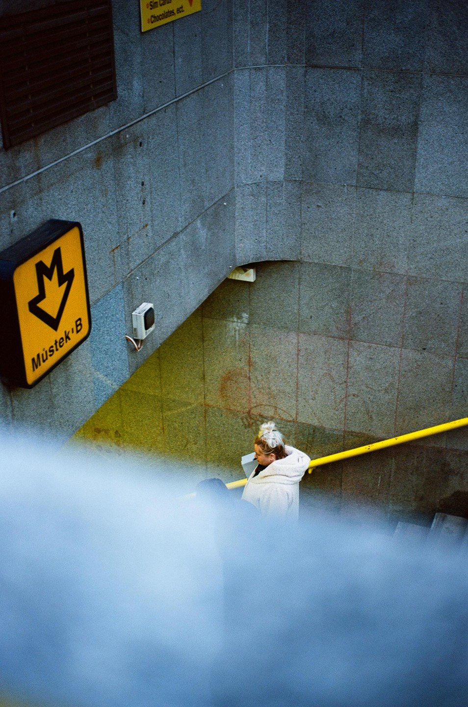 A woman with a bun in her hair descends a subway staircase with a yellow handrail and a yellow sign that reads "Mústek B."