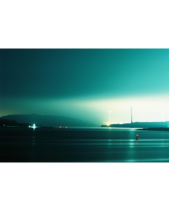 A body of water reflects lights from the shore and buoys under a hazy night sky.