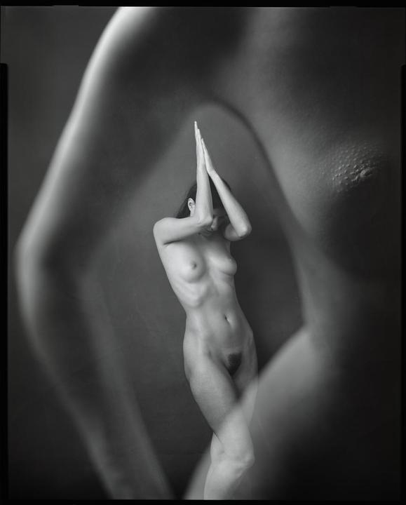 A nude woman stands with her hands together above her head, with a blurred nude body in the foreground.