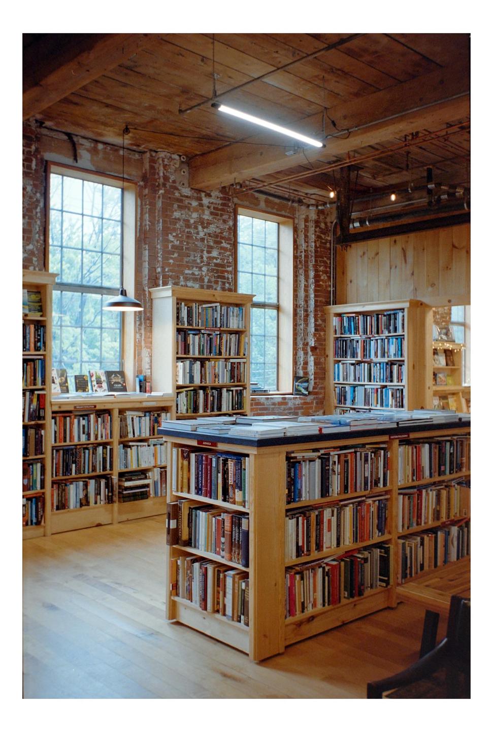 A bookstore interior features tall windows, exposed brick walls, and wooden bookshelves filled with books.