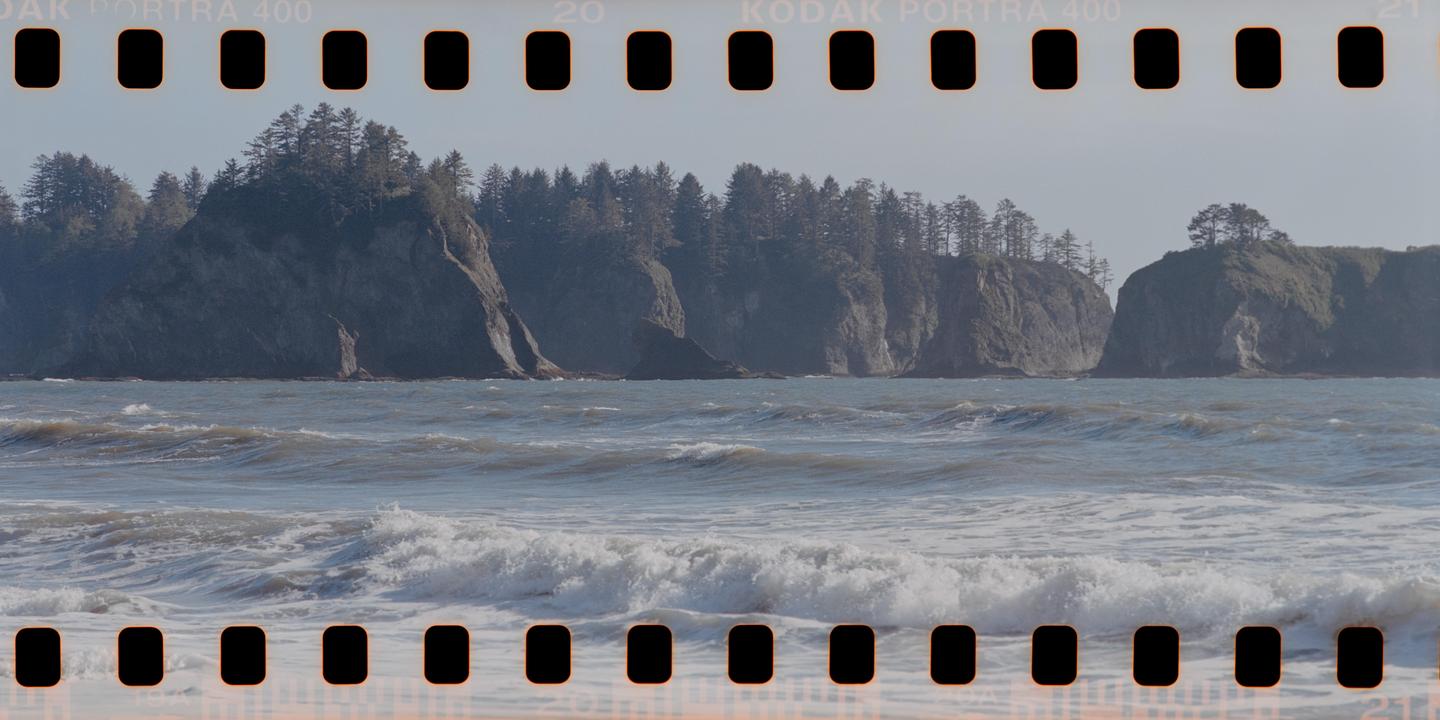 Waves crash on a beach with large, tree-covered cliffs in the background.
