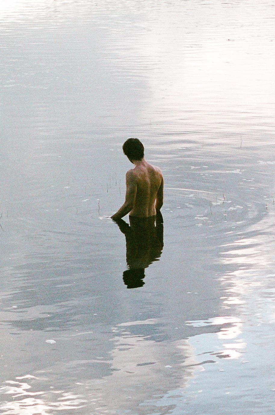 A man stands in still water with his back to the viewer, his reflection visible below him.