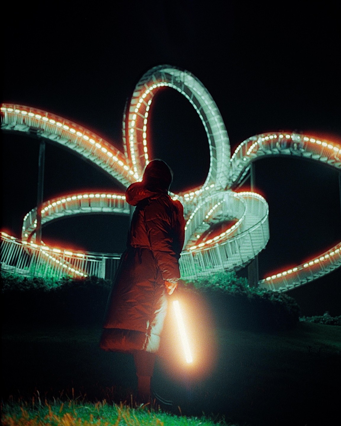 A person in a winter coat holds a glowing light stick while standing in front of a large, illuminated, looping structure at night.