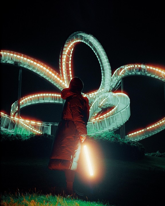 A person in a winter coat holds a glowing light stick while standing in front of a large, illuminated, looping structure at night.