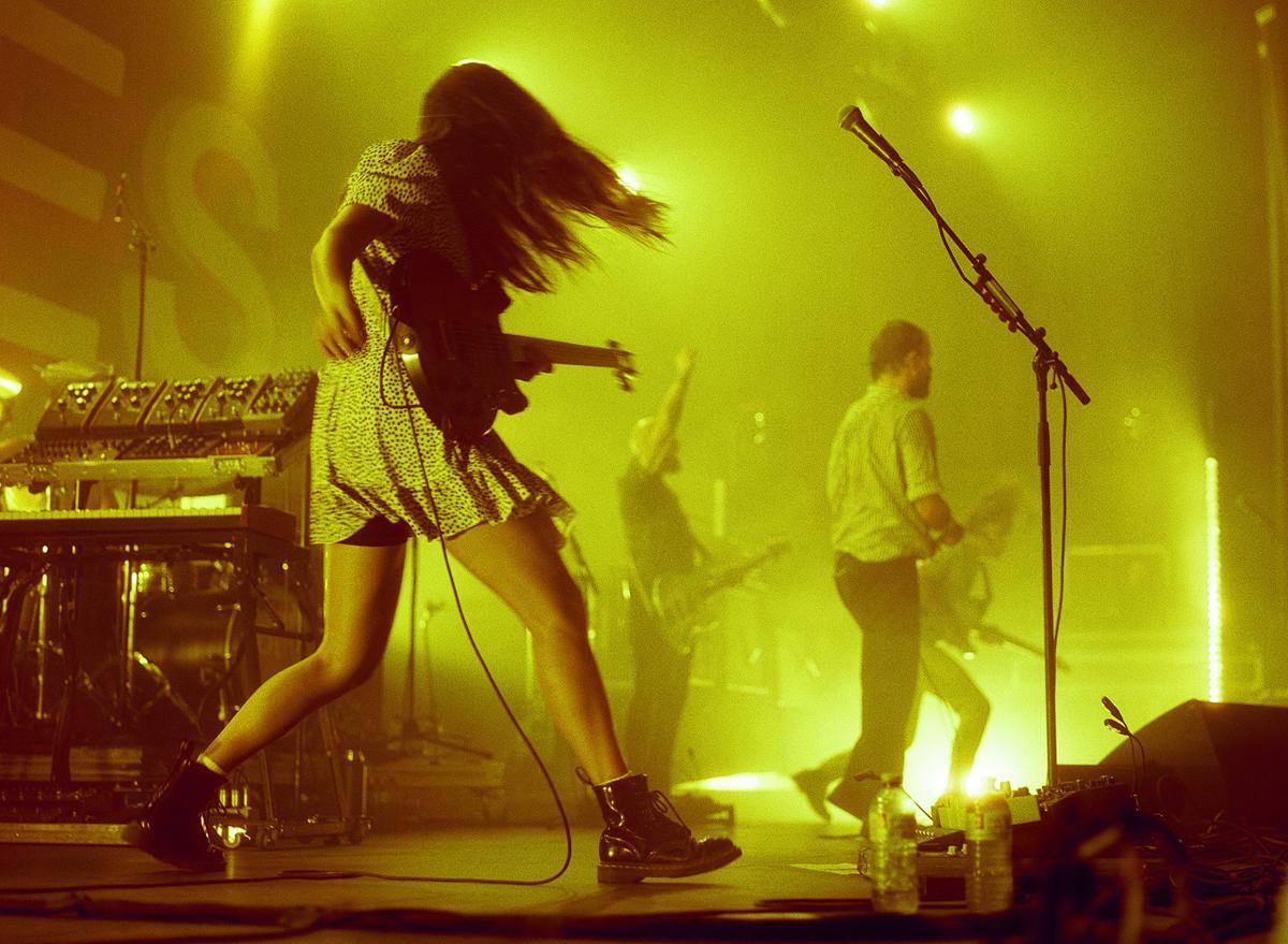 A woman with long hair plays guitar on stage with a band under bright yellow lights.