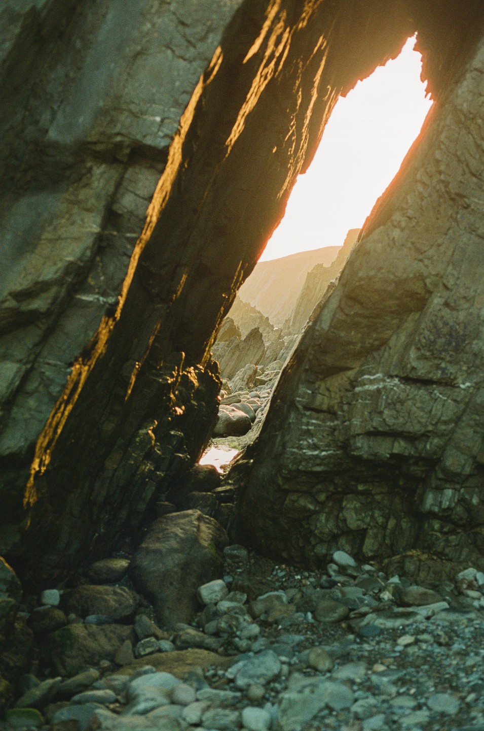 Sunlight shines through a narrow opening between two large rock formations on a rocky beach.
