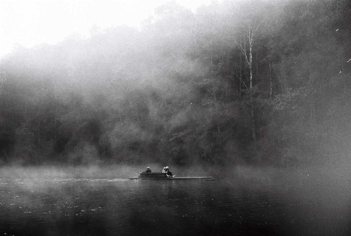 Two people are on a small raft on a misty lake with a forest in the background.