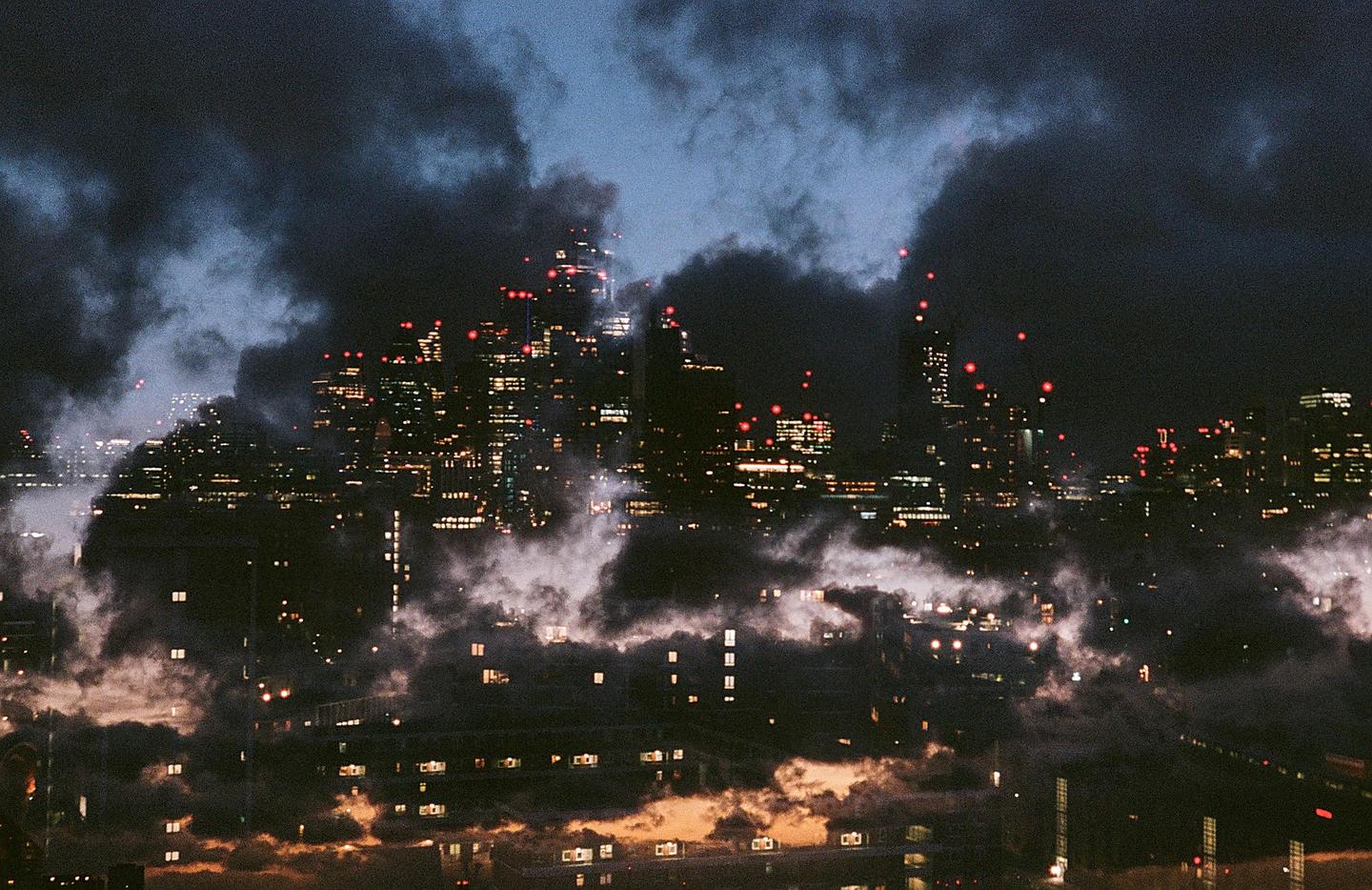 A cityscape at night is shrouded in clouds and fog, with illuminated windows and red lights visible on the buildings.