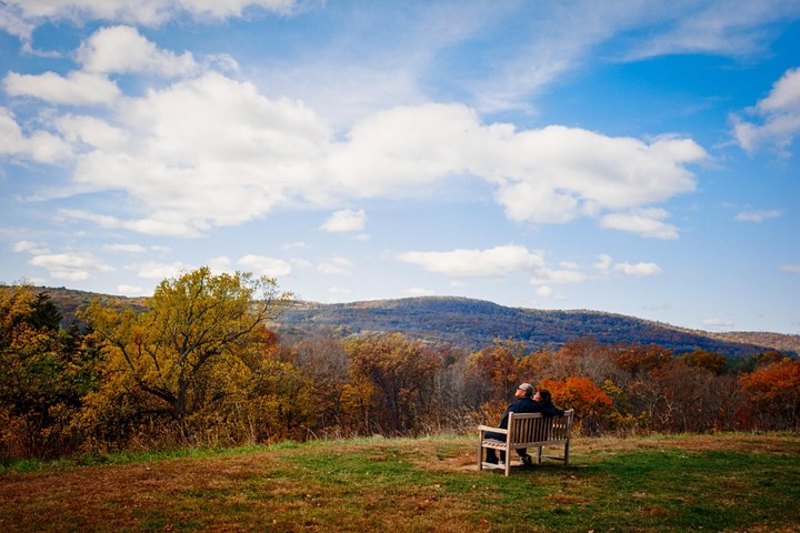 A couple sits on a bench overlooking a valley filled with autumn trees under a blue sky with white clouds.