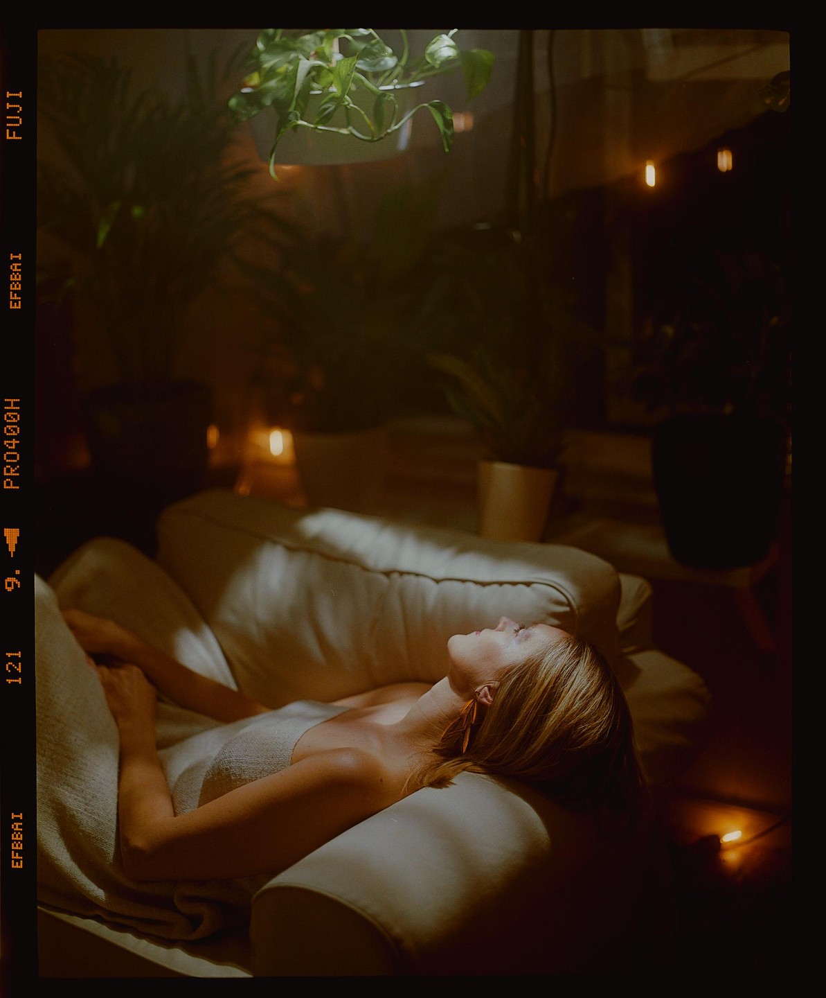 A woman lies on a sofa in a dimly lit room with plants and string lights.
