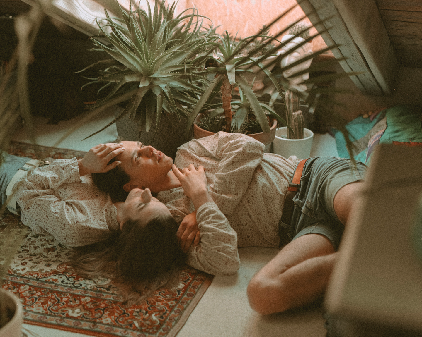 A couple lies on a patterned rug surrounded by potted plants.