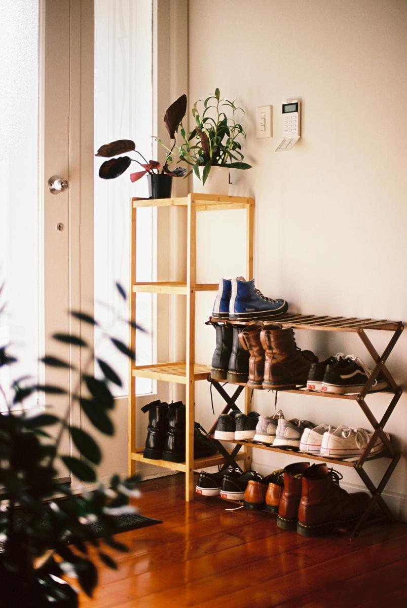 A wooden shoe rack with many pairs of shoes sits next to a door and a plant stand with potted plants.