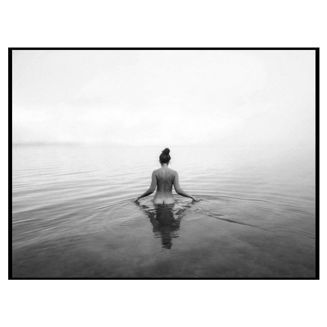 A woman with her back to the viewer stands in calm water under a hazy sky.