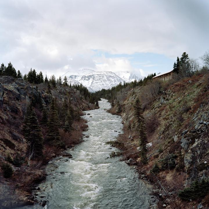 A river flows through a valley with snow-capped mountains in the distance and a building on the right hillside.