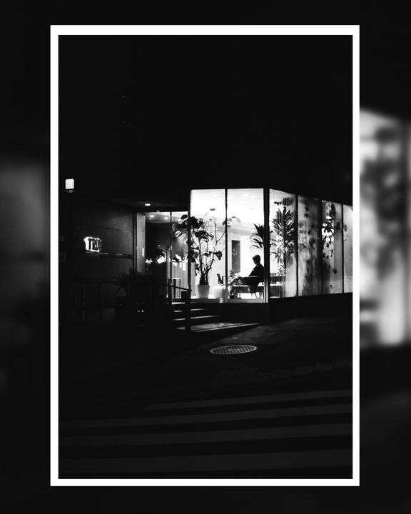 A person sits inside a brightly lit building at night, with a crosswalk visible in the foreground.