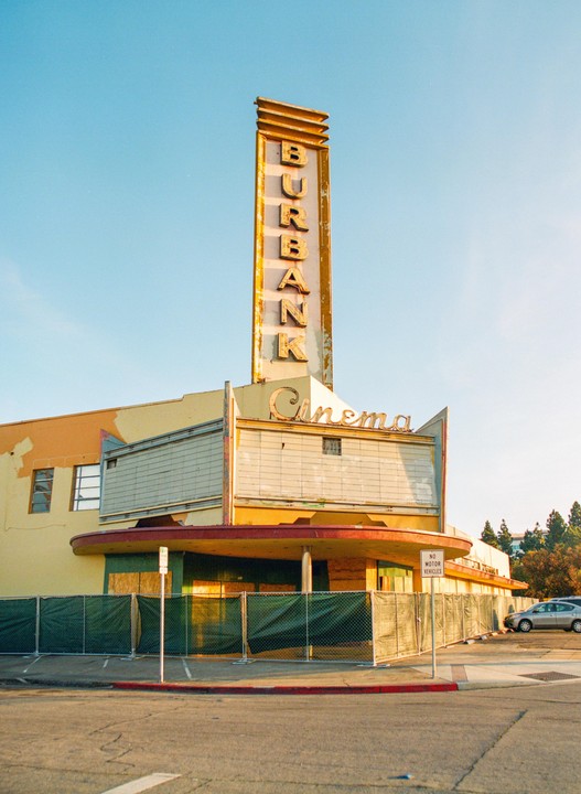 An old cinema with a tall sign that says "Burbank" and "Cinema" is fenced off on a sunny day.
