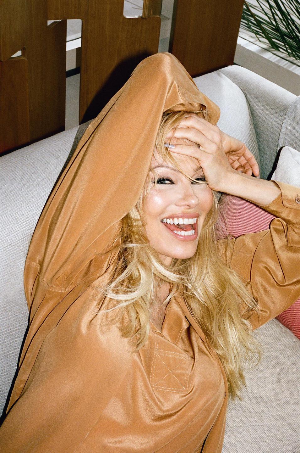 A woman with long blonde hair laughs while covering her forehead with her hand.