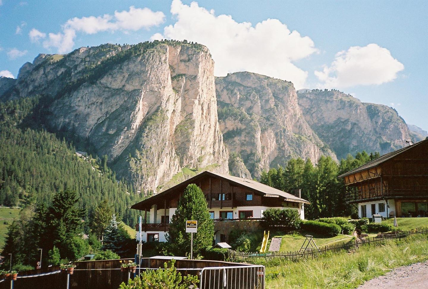 A large house with a playground in the yard is nestled at the base of a towering, rocky mountain range covered in trees.