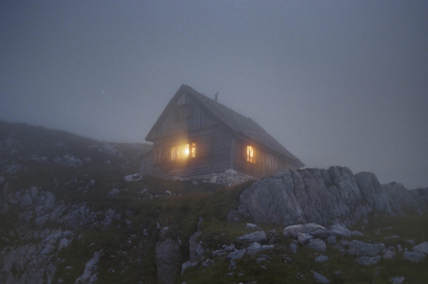 A wooden lodge with illuminated windows sits on a rocky, grassy hillside shrouded in fog.